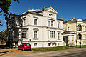 Die weiße Villa weist Elemente der Neorenaissance, wie runde Giebelaufsätze und Pilaster auf. Davor parkt ein rotes Auto. 
