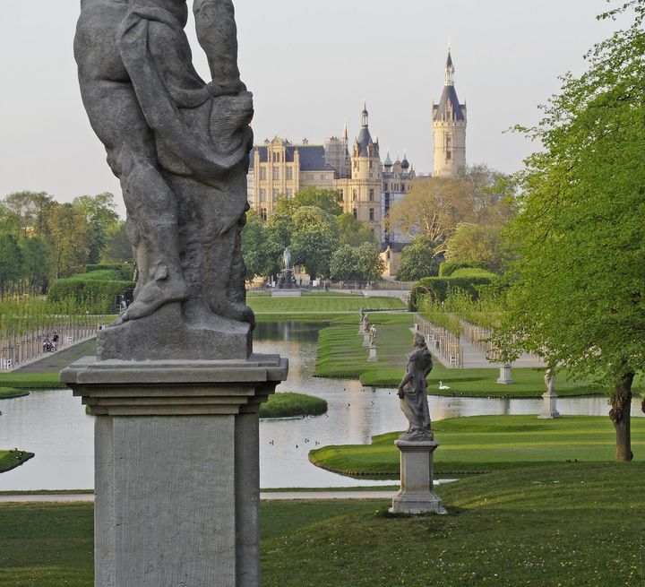 Im Hintergrund einer besockelten Sandsteinstatue befinden sich die Kanäle des Burggartens und das Residenzschloss.