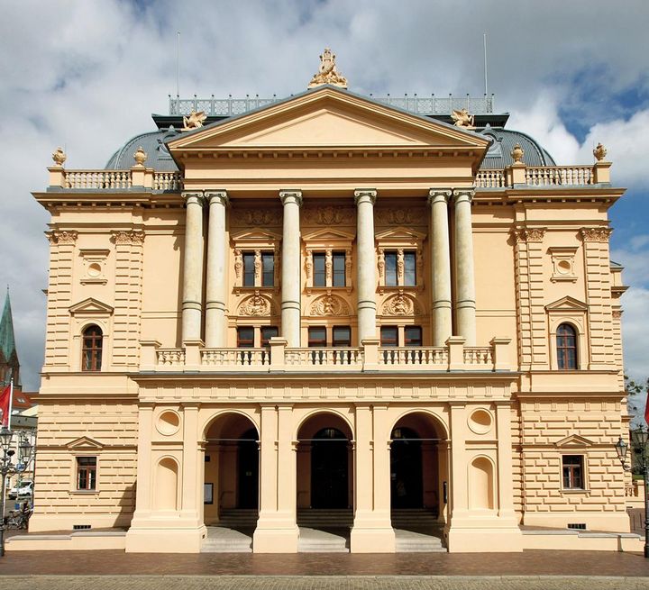 Die Frontansicht des Staatstheaters zeigt die Fassade im Stil der Neorenaissance mit verglaster Loggia mit Terrasse und Vorplatz.