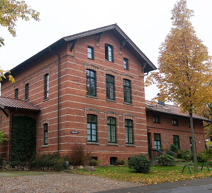 Die Aufnahme zeigt einen kleinen einstöckigen Anbau linksseitig am Wohngebäude. Das gesamte Höfgärtner-Etablissement ist aus rotem Backstein mit hellen Ziegelbändern. Die Fensterrahmen sind dunkelgrün.