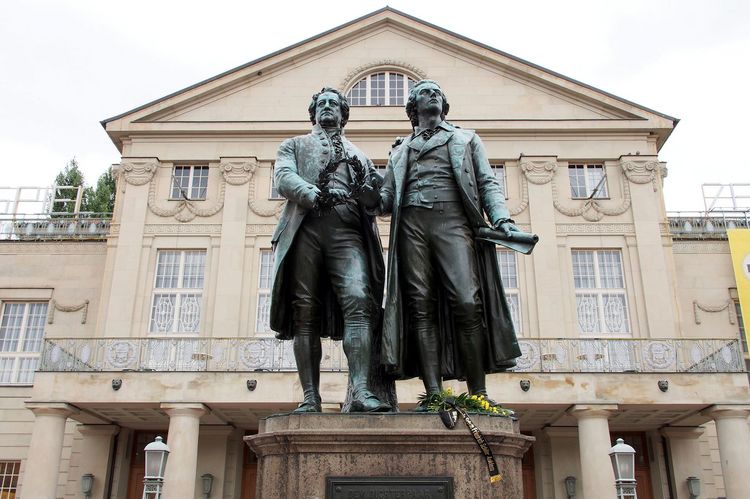 Die bronzene Doppelstatue von Goethe und Schiller auf einem Steinsockel vor dem neoklassizistischem Bau des Deutschen Nationaltheaters. Goethes linke Hand erfasst die Schulter Schillers, während er ihm mit der rechten den Lorbeerkranz reicht. Schiller hält in der linken Hand eine Schriftrolle, während die rechte nach dem Kranz greift.
