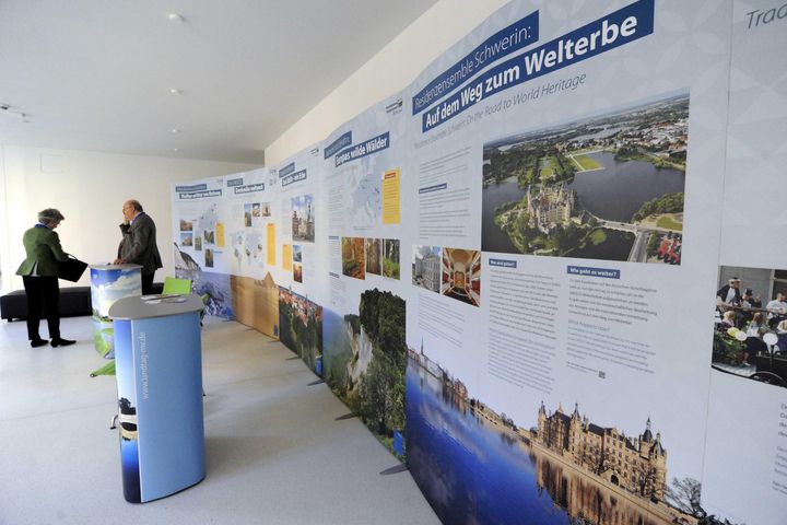 Ein deckenhohes Stehbanner zieht sich entlang einer Wand und zeigt Artikel und Bilder zum Residenzenseble Schwerin. Im Hintergrund unterhalten sich zwei Personen an einem der zwei Podiumstische.