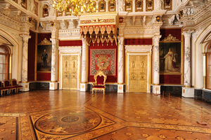 Die Innenansicht des Thronsaales zeigt den Thron zwischen goldenen Türen, Gemälden, Säulen und prunkvollen Wandornamenten.